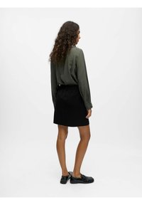 Grüne Bluse mit langen Ärmeln und geknöpften Manschetten, kombiniert mit einem schwarzen knielangen Rock. Modell trägt schwarze Slipper. Glatte Textur und lässige Passform.