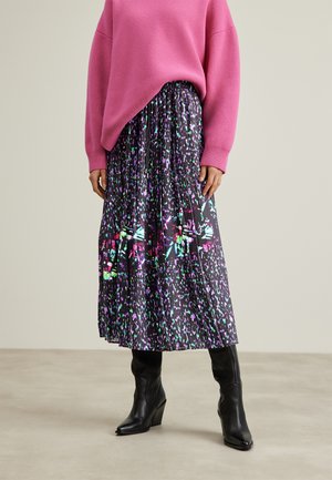 Kvinde iført en sort plissee-midisnørk med farverige abstrakte mønstre, en løs lyserød sweater og sorte kilehælede støvler.