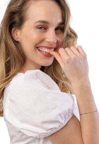 Portret van een persoon met lang, golvend, lichtbruin haar, gekleed in een witte top met pofmouwen, die glimlacht en zijn vingers op de lippen rust, met een stralende huid.