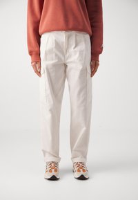 Pantalons utilitaires blancs avec poches latérales, associés à un sweat-shirt corail. Le mannequin porte des baskets beiges avec des accents orange. Texture lisse dans l'ensemble.