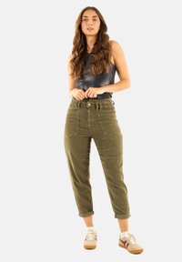 Pantalones de pana verde oliva de talle alto, con bolsillos frontales y puños enrollados, combinados con una blusa de tirantes metálica negra ajustada y zapatos claros.