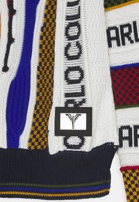 Multicolored knitted sweater featuring white, navy, green, and red sections. Includes a label with a graphic design and text. Textured patterns.