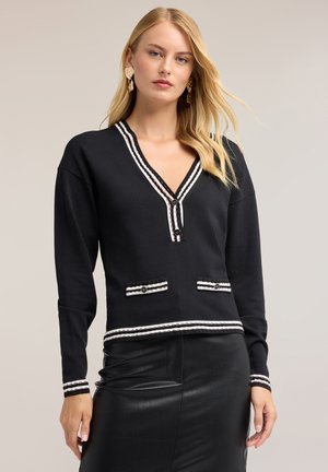 Donna bionda che indossa un maglione nero a maniche lunghe con scollo a V, bordatura bianca e bottoni, abbinato a una gonna in pelle nera e orecchini d'oro.