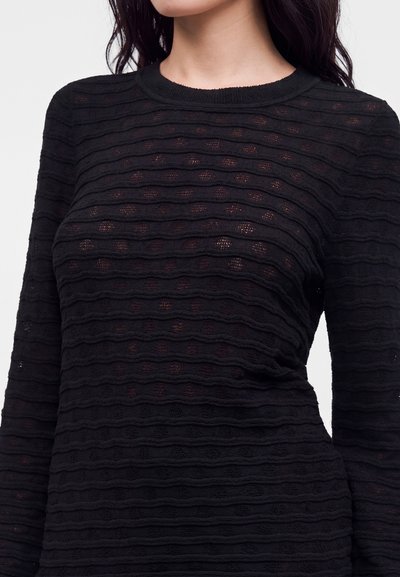 Femme portant un haut noir ajusté à manches longues avec des rayures texturées horizontales et de petites perforations circulaires.