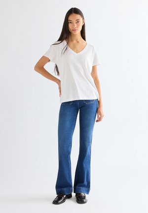 Kvinde stående med den ene hånd på hoften iført hvid V-hals T-shirt, blå vidde jeans og sorte loafers mod en ensfarvet hvid baggrund.