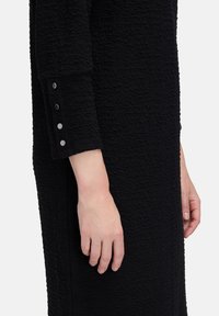 Schwarzes Kleid aus strukturiertem Stoff, tailliertem Schnitt und langen Ärmeln mit drei Metallknöpfen an den Manschetten. Schlichtes und modernes Design.