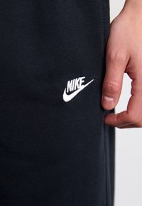 Pantalones de chándal de algodón negros con un logotipo de Nike bordado en blanco en el lado izquierdo. La tela es suave y tiene un ligero brillo.
