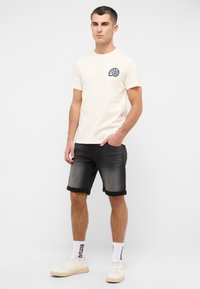 Męska kremowa koszulka bawełniana z granatowym logo graficznym na piersi, w zestawieniu z czarnymi szortami dżinsowymi i białymi trampkami. Prosty, casualowy strój.