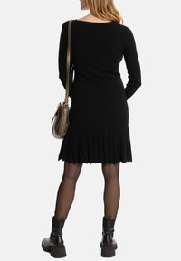 Schwarzes geripptes Kleid mit einem gefältelten Saum, langen Ärmeln, getragen mit schwarzen Stiefeletten und einer metallischen Umhängetasche. Glatte Textur, taillierter Schnitt.