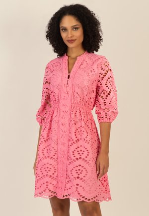 Frau mit lockigem Haar, die ein rosa Midikleid mit Lochmuster, 3/4-Ärmeln und tailliertem Bund trägt, vor einem einfarbigen Hintergrund steht.