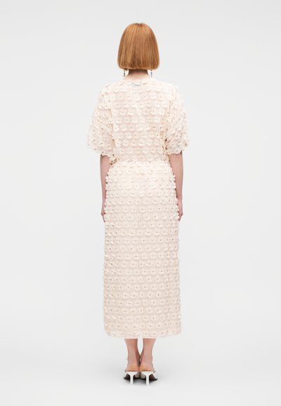 Une longue robe crème avec un motif floral texturé, une taille ajustée, des manches courtes bouffantes et un ourlet droit, portée avec des talons ouverts.