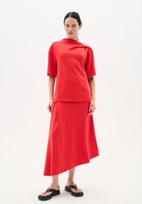 Röd, elastisk klädsel som har en kortärmad topp med asymmetrisk design och en lång, flödande kjol. Svarta platta sandaler kompletterar looken.