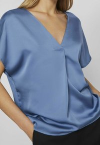 Blouse en satin bleu avec un décolleté en V, des manches courtes et un discret détail de fronces. Texture douce et coupe décontractée.