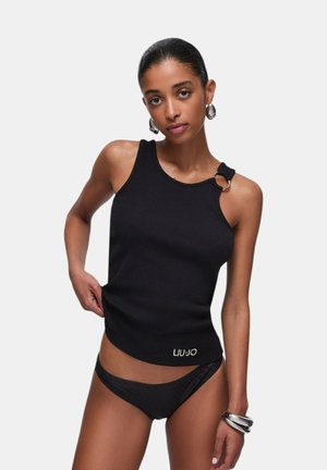 Mujer con un top negro sin mangas con detalle de anillo metálico y braguitas de bikini negras a juego sobre un fondo gris liso.