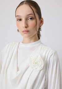 Top bianco con tessuto drappeggiato, caratterizzato da un accento floreale sulla spalla. Texture liscia e scollatura alta, con un fit rilassato.