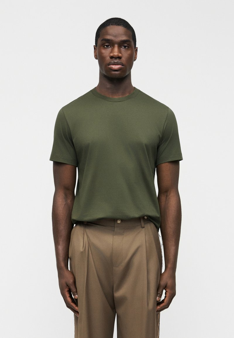 Άνδρας που στέκεται με το πρόσωπο προς τα εμπρός φορώντας μια ελιάς κοντομάνικη μπλούζα και ψηλόμεσα καφέ πλισέ παντελόνια.