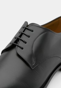 Black leather dress shoe with a smooth finish, featuring a stitched flap and five-eyelet lacing system, emphasizing sleek design and structure.