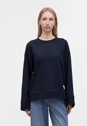 Vrouw met steil blond haar die een loszittend zwart shirt met lange mouwen en een blauwe spijkerbroek draagt, staand tegen een effen witte achtergrond.