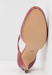 Chaussure rouge à talon haut vue de la semelle, avec un bout pointu, un talon fin et une bride à la cheville sur fond blanc.