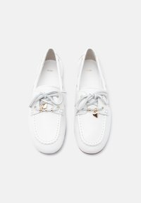 Par de mocasines de piel blanca con cordones grises y pequeños encantos con forma de corazón dorado sobre un fondo blanco.
