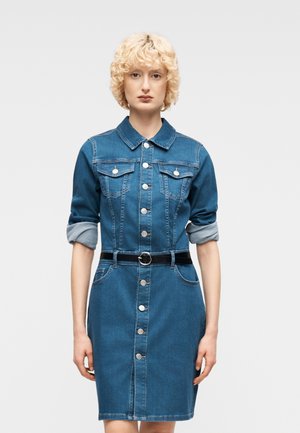 Femme aux cheveux blonds courts et bouclés, portant une robe en denim bleu boutonnée avec manches retroussées et ceinture noire, debout devant un fond uni.