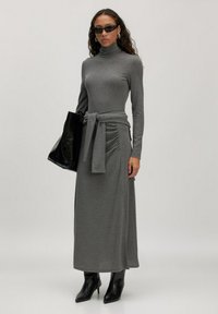 Grijs langslijfjurk met een col, een ceintuur op de taille en geplooide zijdetails. Gecombineerd met zwarte zonnebril en een zwarte tas.