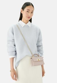 Jasnoniebieski sweter z miękkiego materiału, z prążkowanym dekoltem. Trzyma małą różową torebkę z złotymi akcentami, noszoną na tle białej spódnicy.