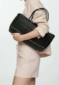 Czarna pleciona torebka z dwoma plecionymi uchwytami, o prostokątnym kształcie i teksturowanej powierzchni, trzymana na tle jasnego materiałowego stroju.