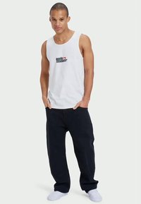 White cotton tank top featuring a logo in black and red, paired with loose-fitting navy pants and white footwear. Simple, casual design.