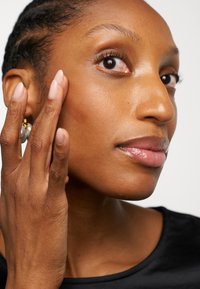 Close-up of a person’s face, highlighting smooth, radiant skin, light brown tone, natural makeup, and large pearl earrings.