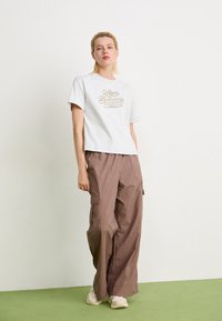 T-shirt en coton gris avec le logo "New Balance", associé à un pantalon cargo beige clair avec des poches latérales et des baskets de couleur neutre.