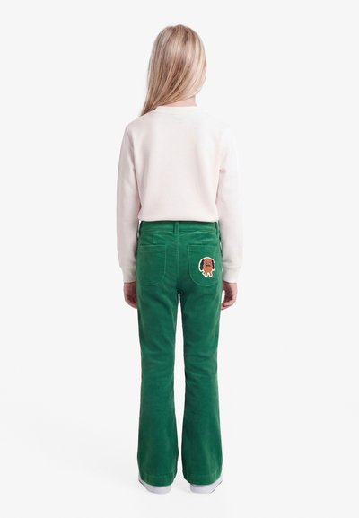 Παιδί με μακριά ξανθά μαλλιά που φοράει λευκή μπλούζα με μακρύ μανίκι και πράσινο παντελόνι με σχέδιο σκύλου στην πίσω τσέπη, στέκεται με την πλάτη στραμμένη.