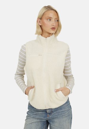 Vrouw met een blond bobkapsel, die een crèmekleurig fleecevest met rits draagt over een beige-wit gestreepte lange mouwen shirt en een blauwe jeans.