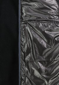 Nahaufnahme von schwarzem Stoff mit glattem, glänzendem synthetischem Textil auf der rechten Seite und mattem, fleeceähnlichem Material auf der linken Seite.