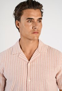 Jasnoróżowa koszula z krótkim rękawem, z pionowymi białymi paskami, kołnierzykiem i dwoma guzikami, wykonana z tkaniny o teksturowanym wzorze.