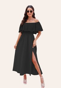 Abito maxi nero senza spalline con maniche a volant, vita elastica, spacco laterale e tessuto leggero; abbinato a sandali bianchi con cinturini.