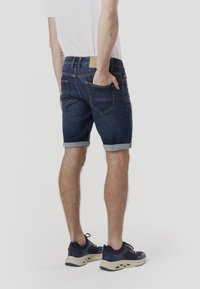 Short en jean avec un lavage bleu foncé, ourlet retroussé et poches arrière. Associé à des baskets bleu marine, le mannequin pose devant un fond neutre.