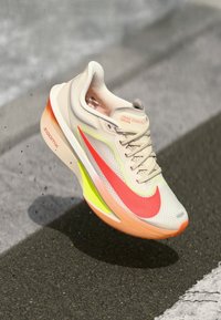 Beige en witte sneaker met roze en neon groene accenten, bovenwerk van mesh, textuurzool en een opvallende rode Nike swoosh.