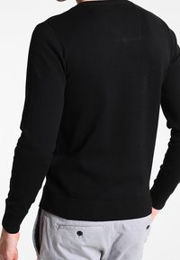 Svart crew-neck tröja med långa ärmar, ribbade muddar och nederkant, slät textur och minimal design. Bakifrån syns inga synliga mönster.