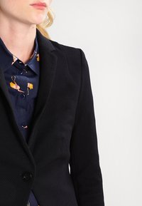 Musta teksturoitu bleiseri istuvalla mallilla, jossa on yksittäinen nappikiinnitys ja lovikääntöpäällinen, puettu tummansinisen kuvioidun paidan päälle.