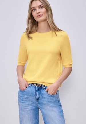 Femme aux cheveux blonds portant un pull à manches courtes jaune et un jean bleu clair avec une ceinture à motifs, les mains dans les poches, debout devant un fond gris.