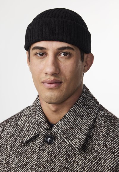 Cappotto a motivo houndstooth marrone e crema con un bottone nero, abbinato a un berretto a maglia nera a coste, primo piano del volto di un modello maschile.