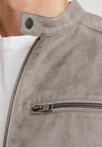 Giacca grigia in faux suede con colletto alto, chiusura con cerniera argentata e tasca sul petto con zip. Texture morbida e design minimalista.