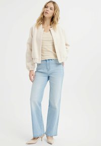 Puchowa kurtka bomber w odcieniu ecru na tle paskowanego beżowego topu, zestawiona z jasnoniebieskimi spodniami z wysokim stanem i beżowymi szpilkami.