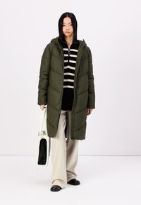 Abrigo largo acolchado en verde oscuro con capucha, que presenta un cierre de cremallera y un acolchado vertical. Combinado con una camiseta de rayas y pantalones anchos de color claro.