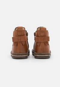 Des bottines en cuir marron présentent un bord supérieur festonné, deux rubans et une semelle texturée avec des coutures contrastées. Fermeture à l'arrière.