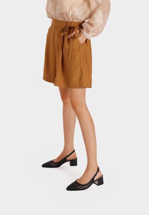 Kobieta w brązowych szortach z wysokim stanem i wiązanym paskiem, przezroczystej białej bluzce oraz czarnych butach na niskim obcasie typu slingback, stojąca na białej podłodze.