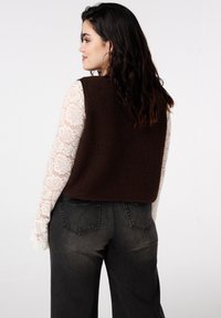 Bruin fleecevest met een gestructureerde afwerking, gedragen over een wit kanten shirt met lange mouwen, gecombineerd met donkergrijze jeans met brede pijpen.