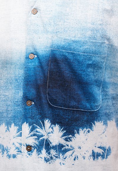 Μπροστινό μέρος πουκάμισου με καφέ κουμπιά, τσέπη με μπλε βαθμονόμηση και λευκό σχέδιο με φοίνικες κατά μήκος του κάτω άκρου.