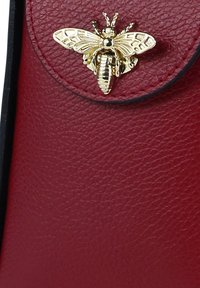 Sac en cuir rouge texturé avec une broche en forme d'abeille dorée détaillée comme fermoir. Le design comprend un rabat arrondi et une bordure noire contrastante.